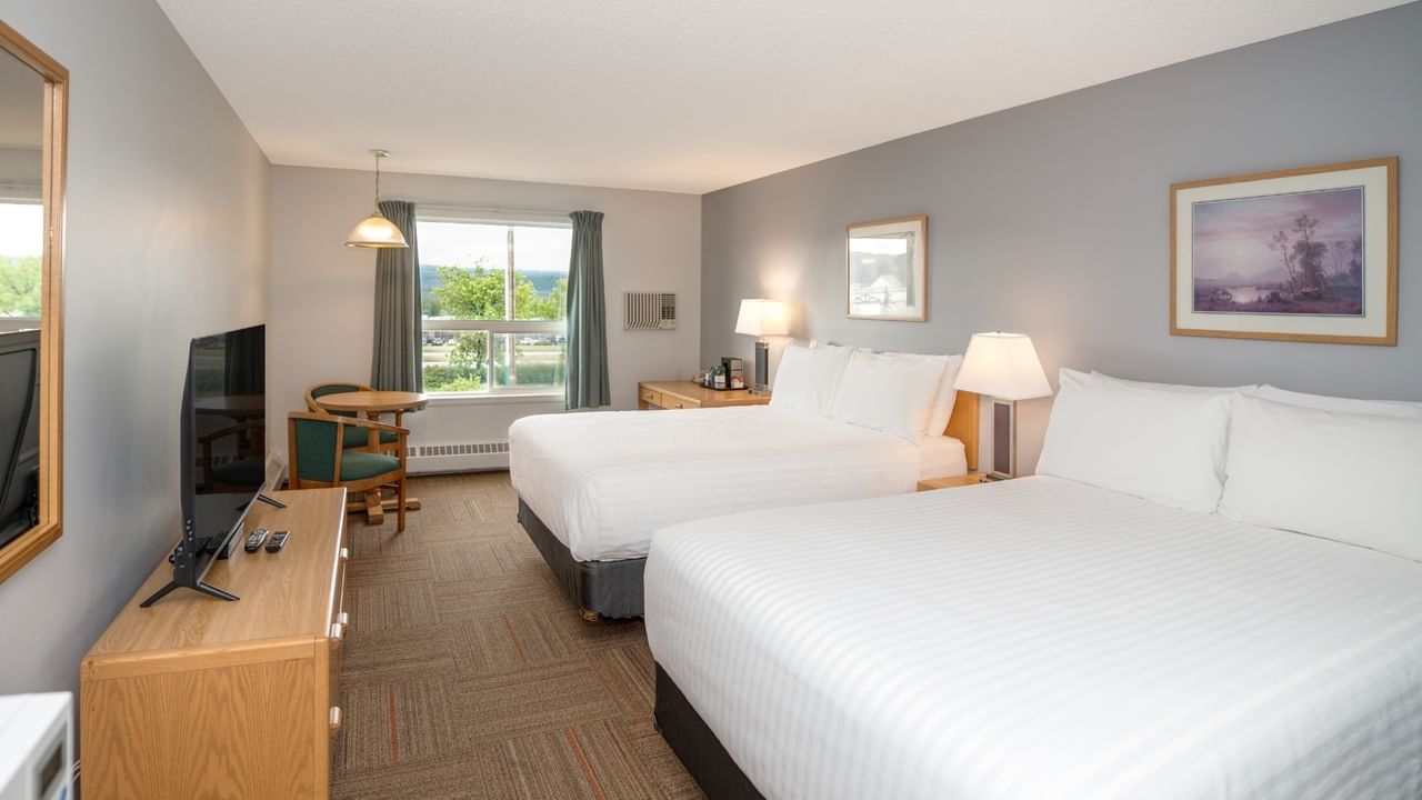 Two beds in a hotel room with nightstands, lamps, TV, table, chairs, and window.