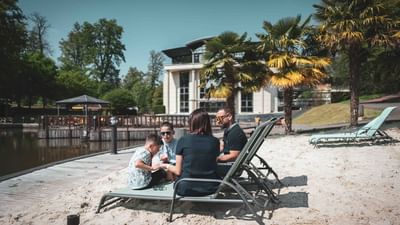 Four people sit on lounge chairs by a sandy shore with palm trees and a modern building.