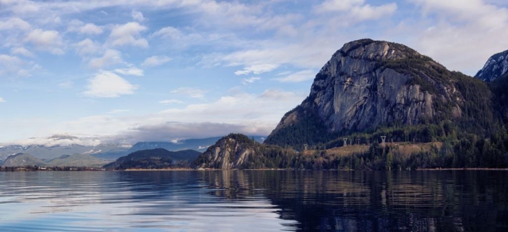 The Stawamus Chief rising above reflective waters in Squamish, framed by blue skies and distant mountain peaks.