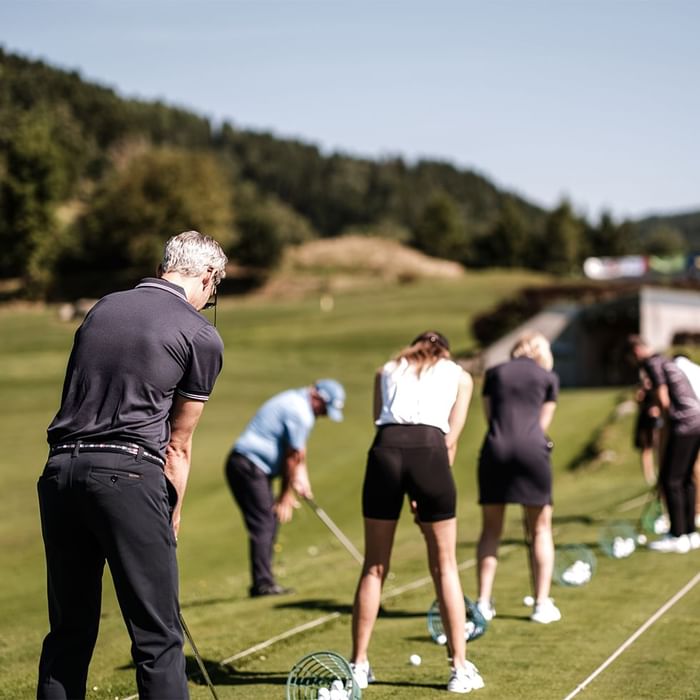 Group of people practicing golf on a green field with trees and hills in the background.