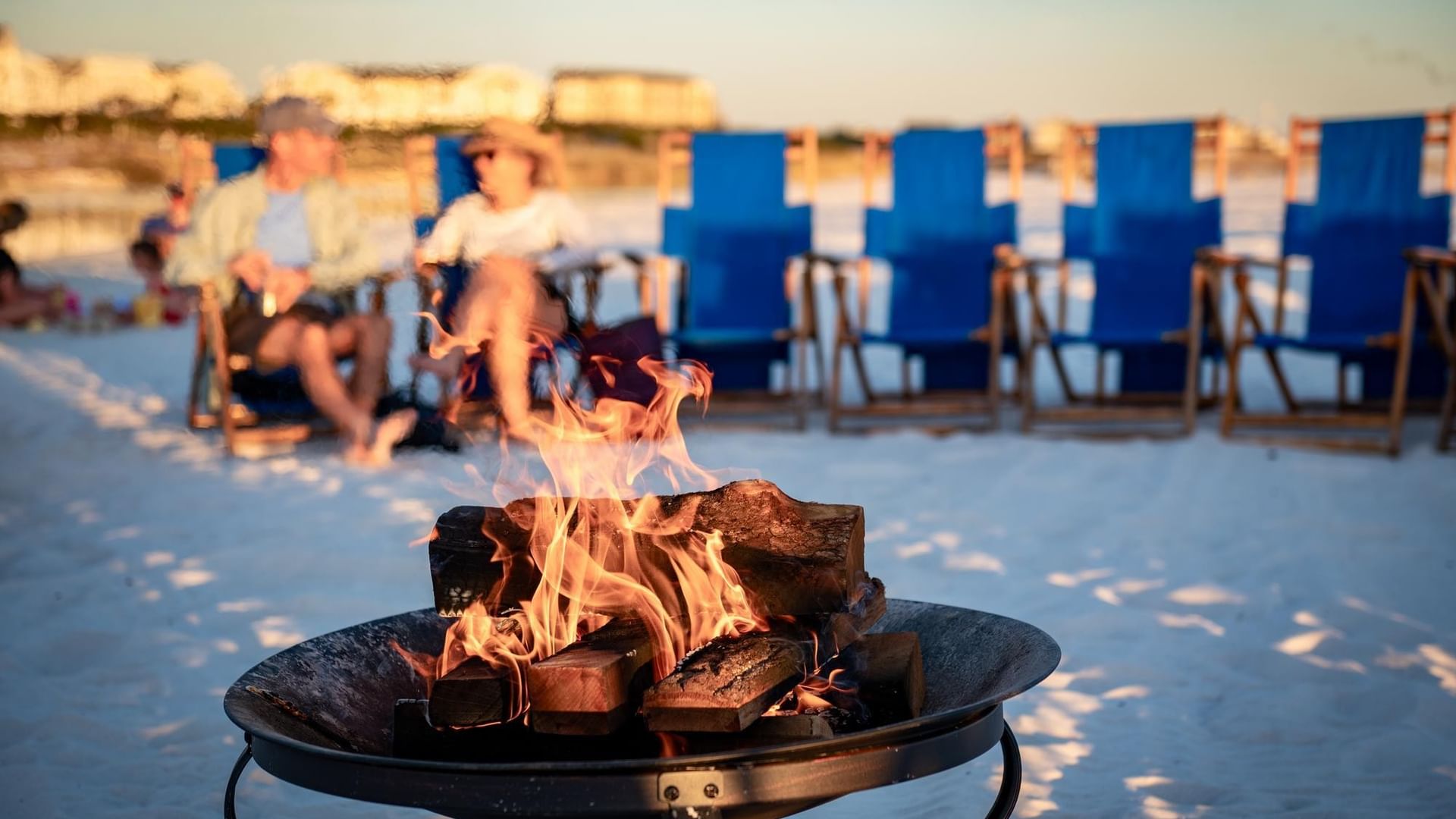 People sit in beach chairs near a fire pit on the sand, with buildings in the background.