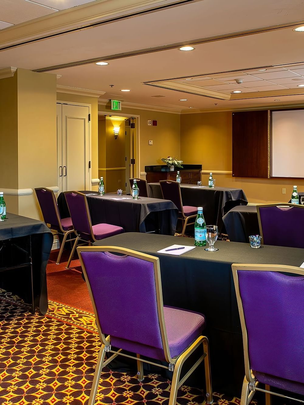 Purple chairs by black tables under a projector screen near a wooden lectern in Tabor I at Warwick Denver