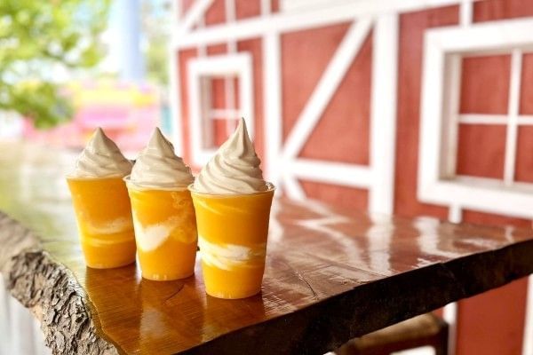Three layered orange and white slushies sit on a wooden table against a red barn backdrop.