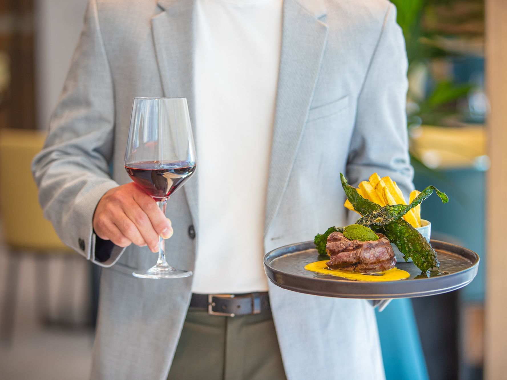 A man in a suit holds a wine glass and a tray with a meat dish.