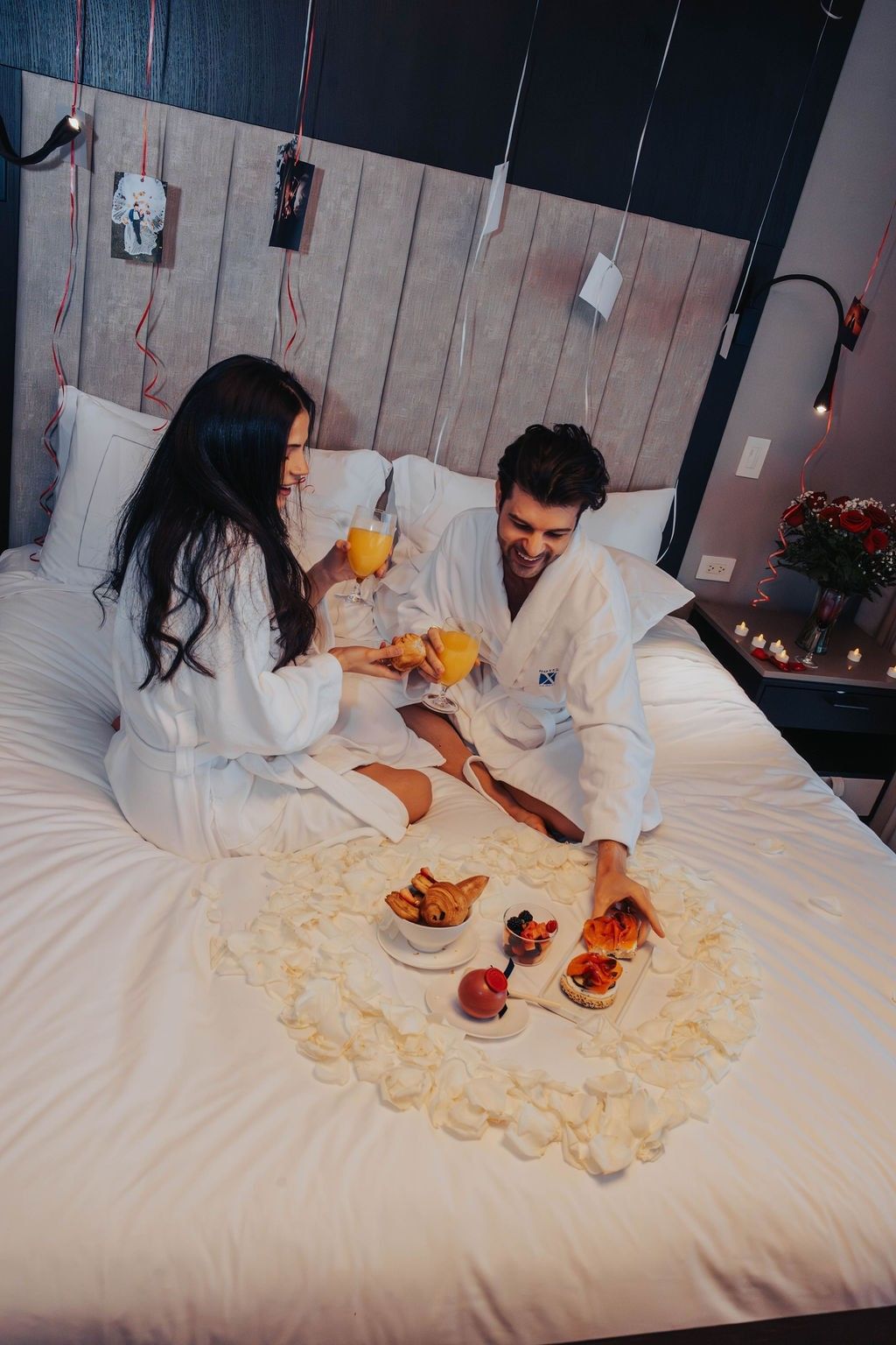 Couple in robes toasting mimosas on a bed surrounded by rose petals and pastries at Hotel X Toronto