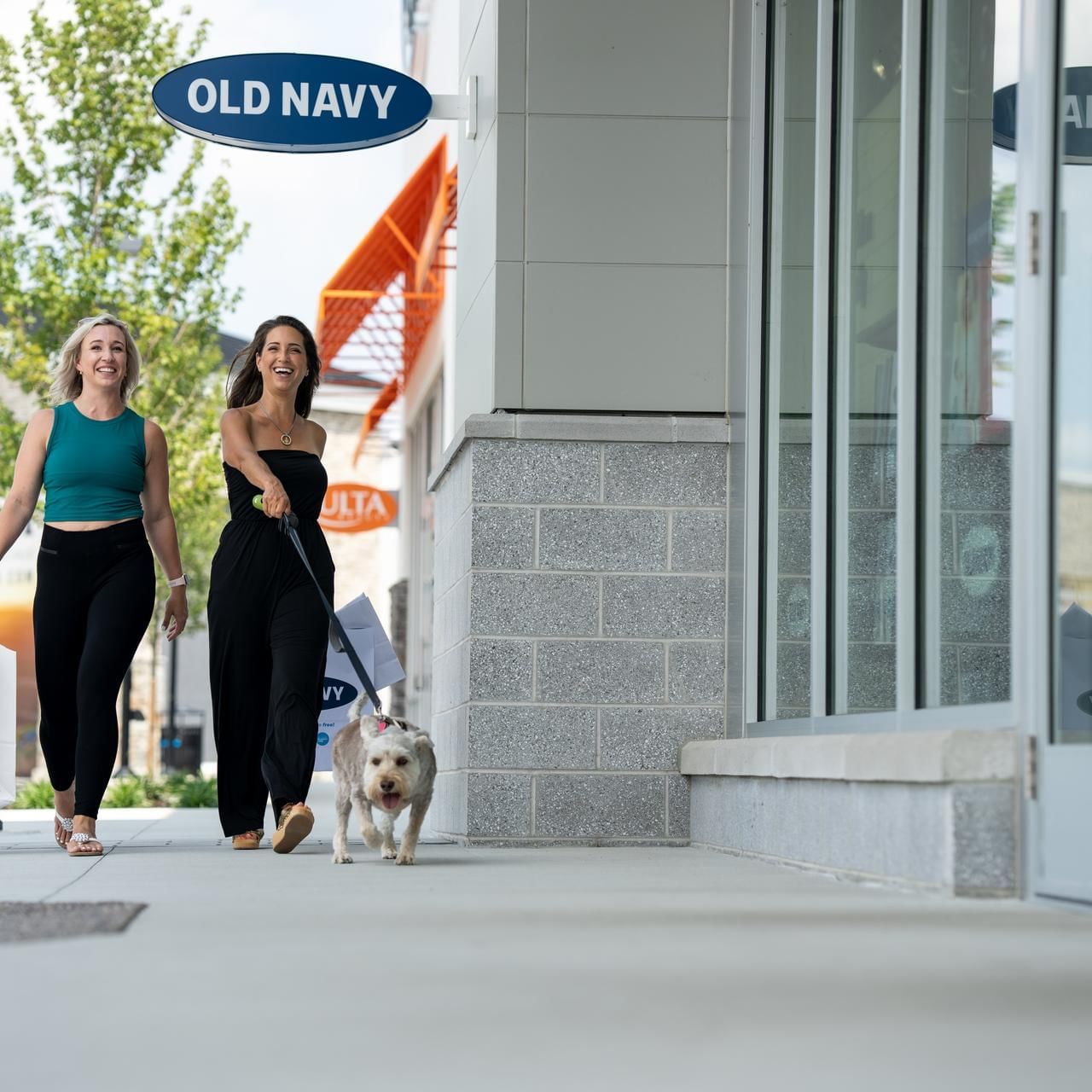 Two ladies strolling with a dog while carrying shopping bags near The Artisan Hotel at Tuscan Village