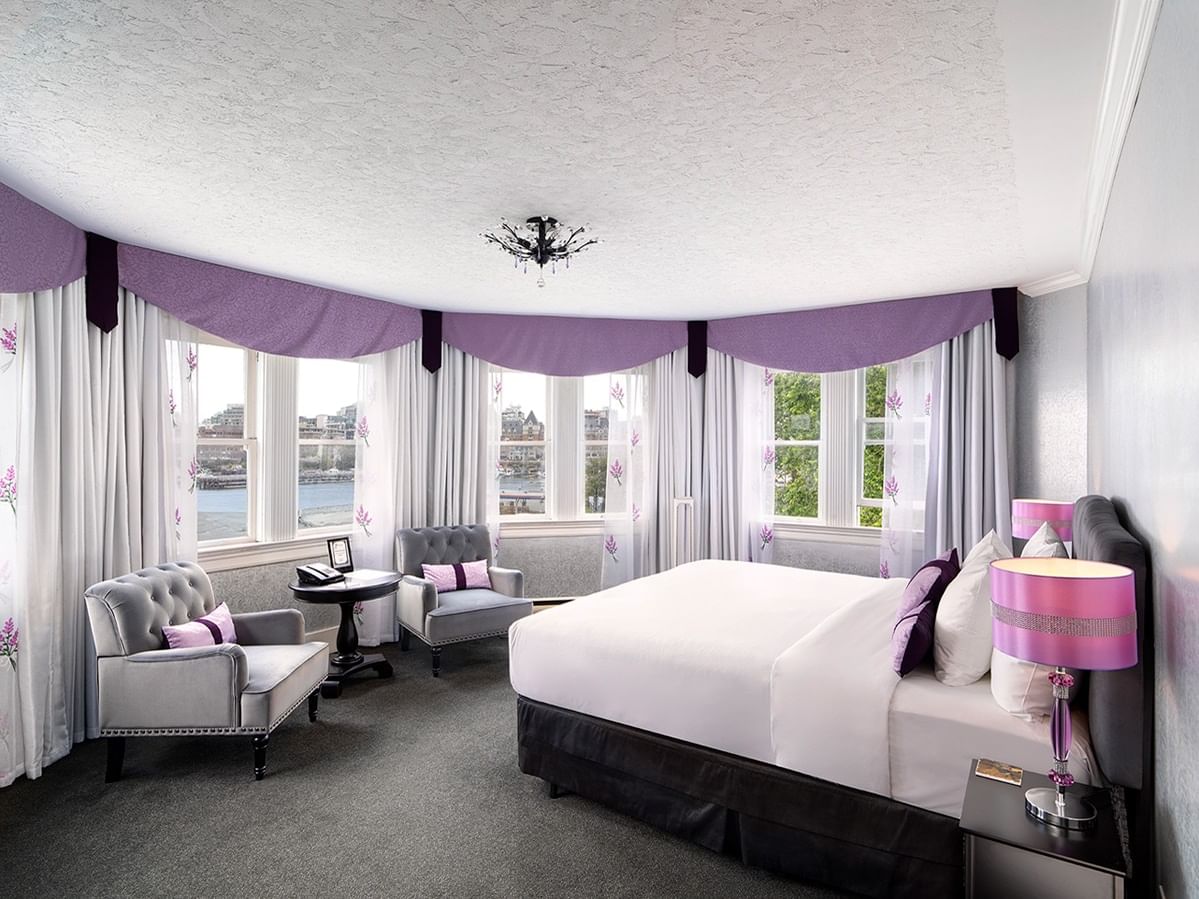A luxurious bedroom with large windows, a king-sized bed, two armchairs, and a round table.