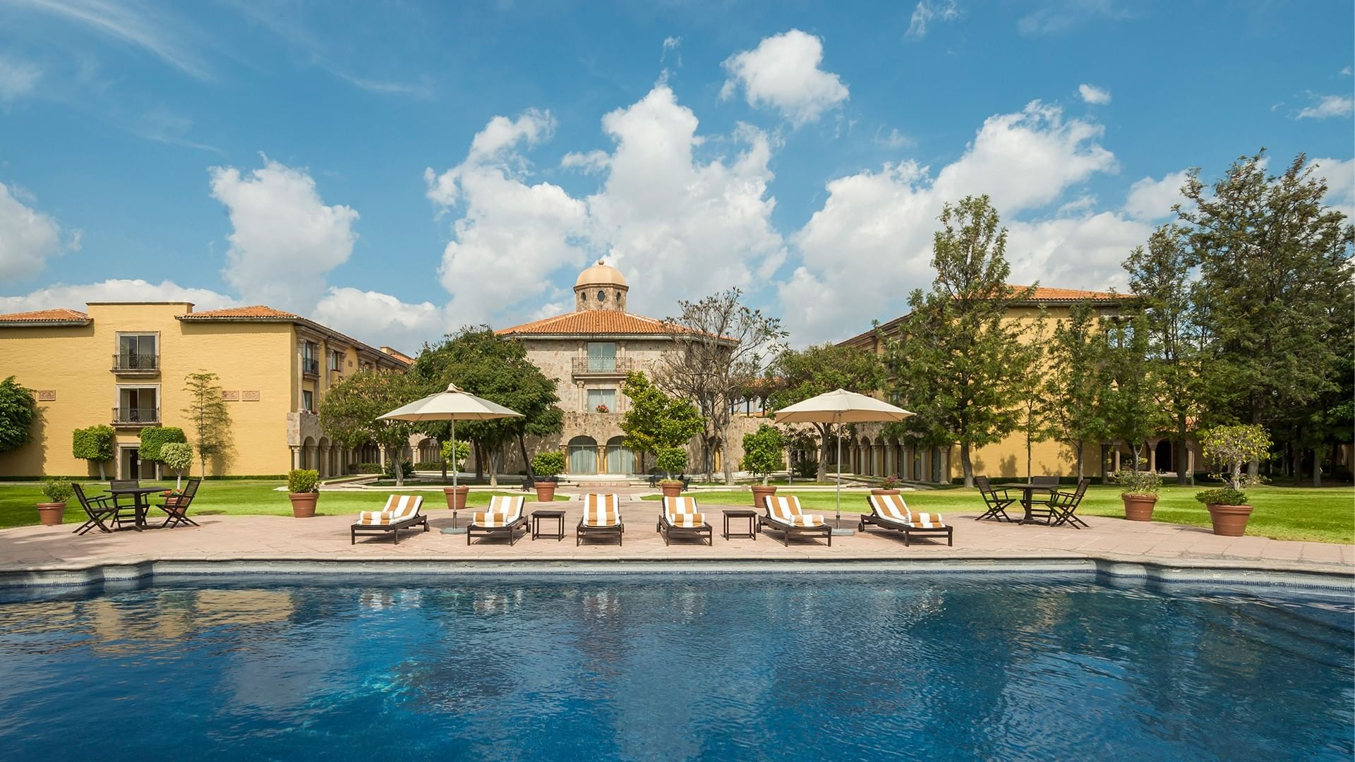 Large blue swimming pool with striped lounge chairs and yellow buildings at Quinta Real Aguascalientes