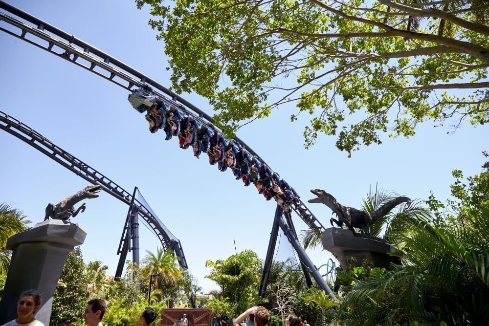 A roller coaster track with upside down riders soaring between two raptors atop columns.  