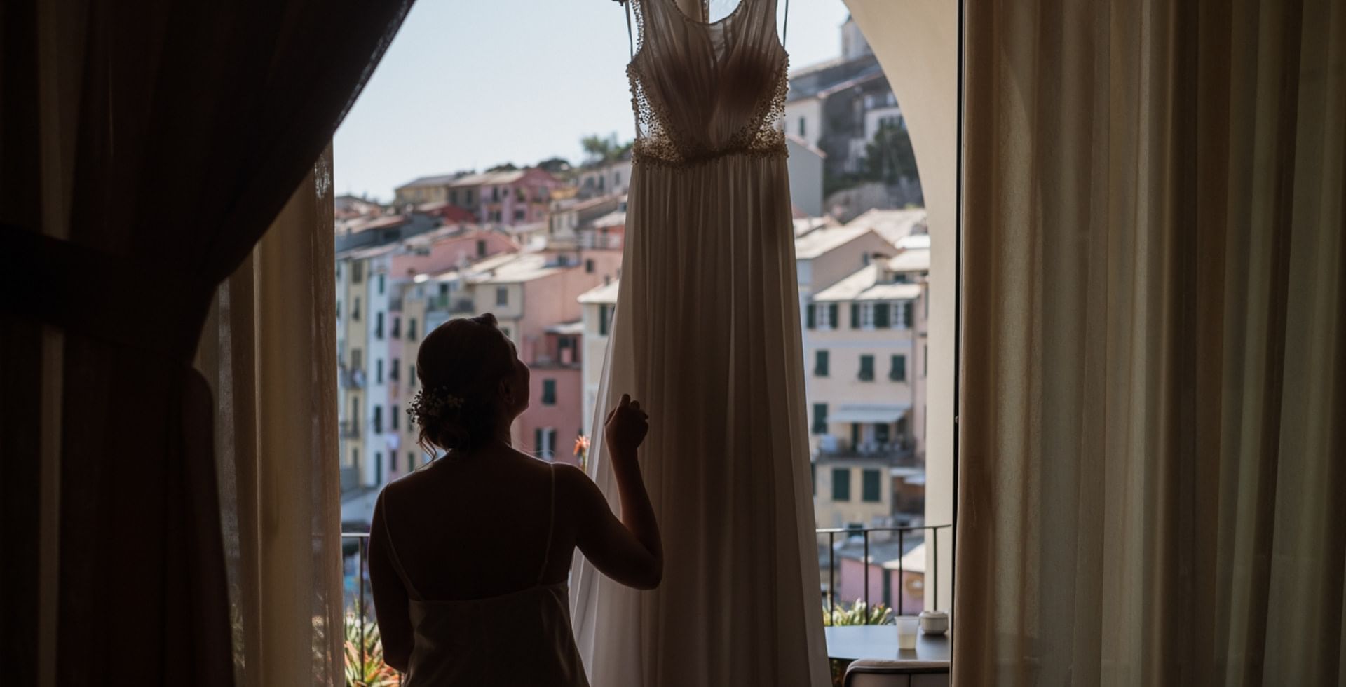 Donna che guarda fuori dalla finestra con abito da sposa appeso e vista su Portovenerere