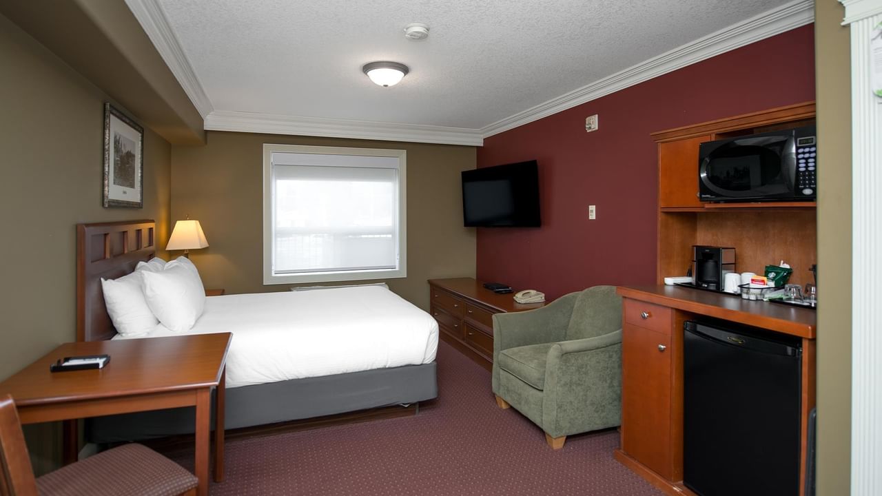 Guest room with queen bed, kitchenette, wall-mounted TV, and olive armchair at Coast Edson Hotel.