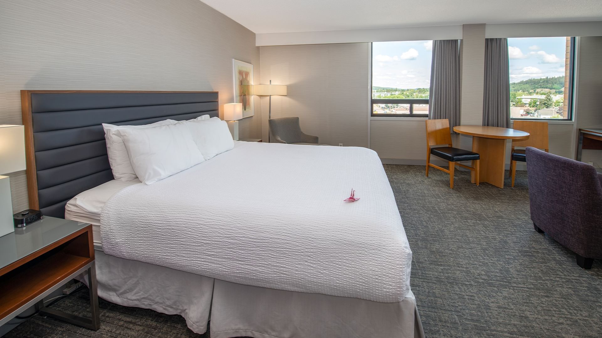 A hotel room with a large bed, nightstand, table, chairs, and windows showing a cityscape.