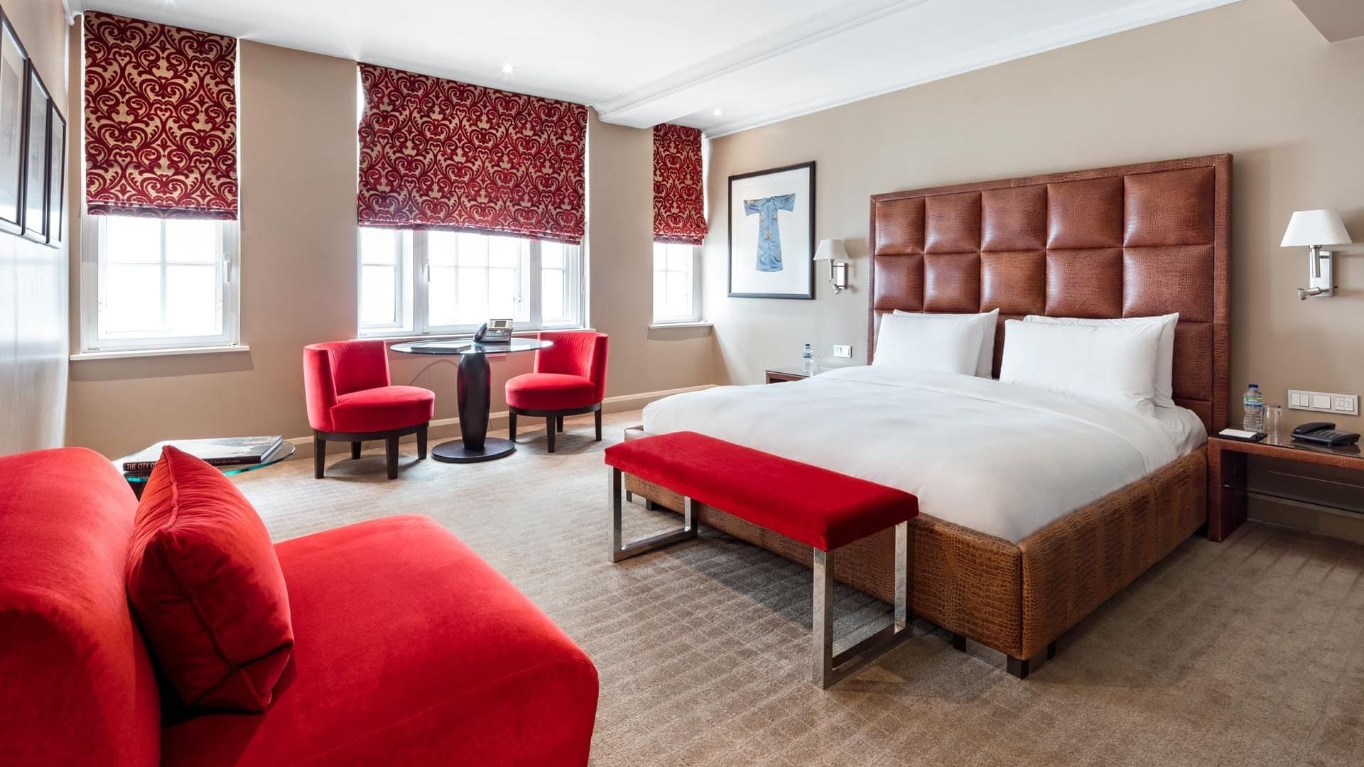 Large bed with padded headboard and red accents in chairs and bench in the Collection Premium Room at The May Fair Hotel