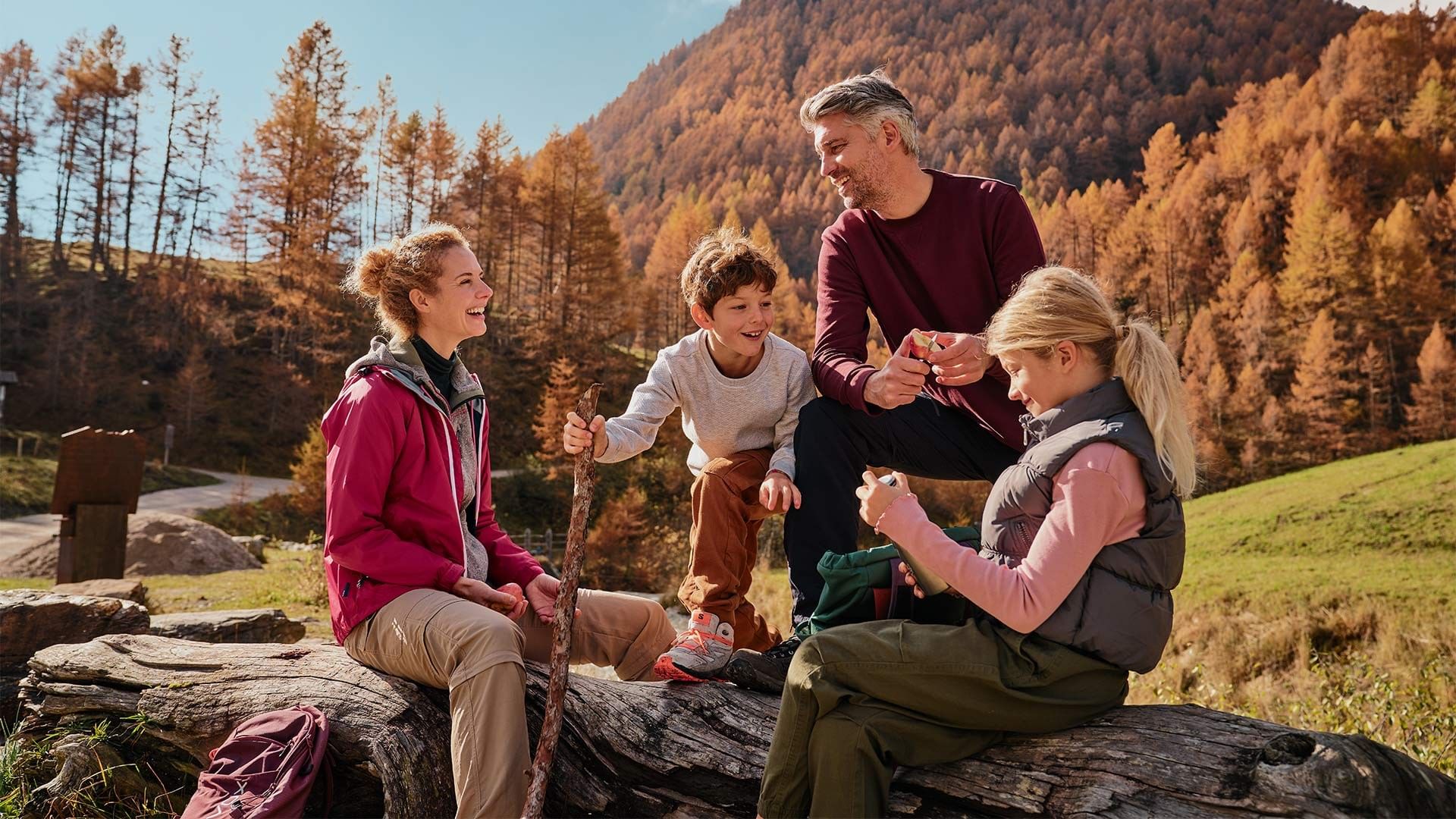 Familie genießt Picknick auf Baumstamm im herbstlichen Bergwald, umgeben von bunten Bäumen.