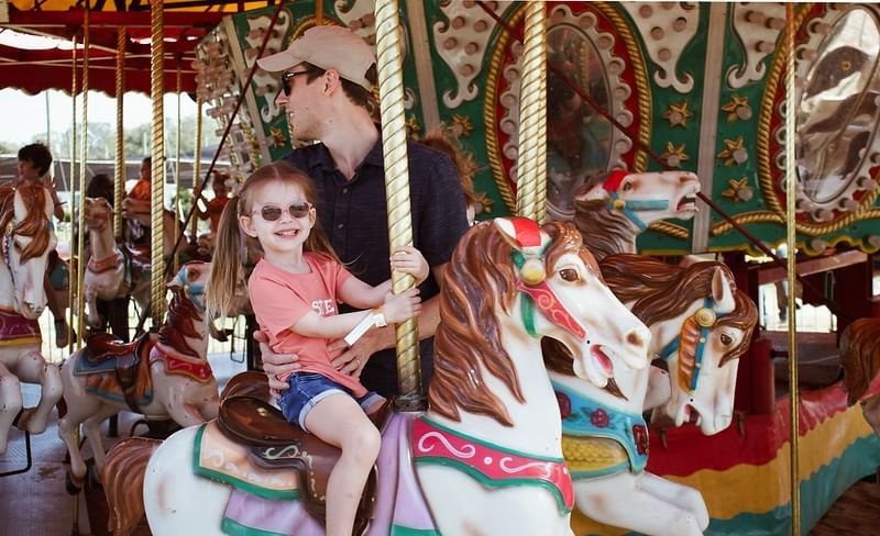 A girl with sunglasses and pigtails rides a carousel horse, smiling at the camera with her dad standing behind her. 