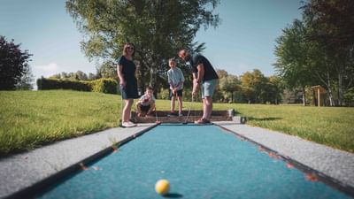 Family of four playing mini golf on a blue mat with trees and grass around.