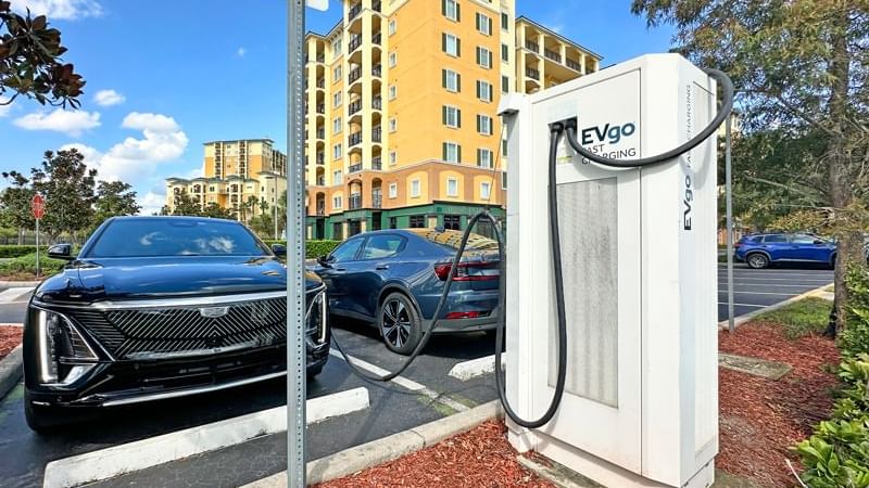 A white EVgo fast charging station with a black cord plugged into a black car in a parking lot with a hotel building in the background.