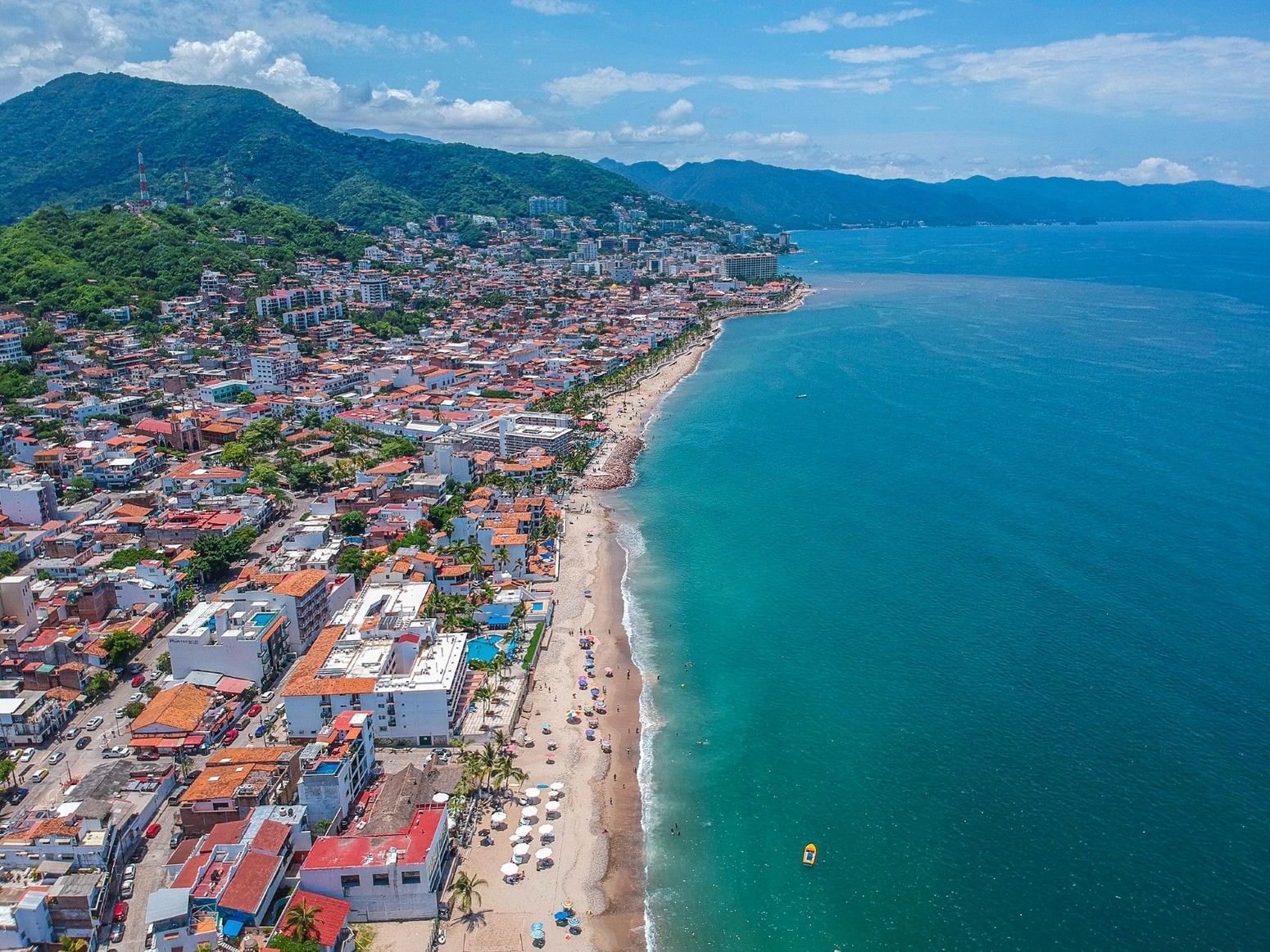 Aerial view of Puerto Vallarta on a sunny day, featuring the Pacific coastline, beach umbrellas along the shore, oceanfront buildings, and lush green mountains in the background.