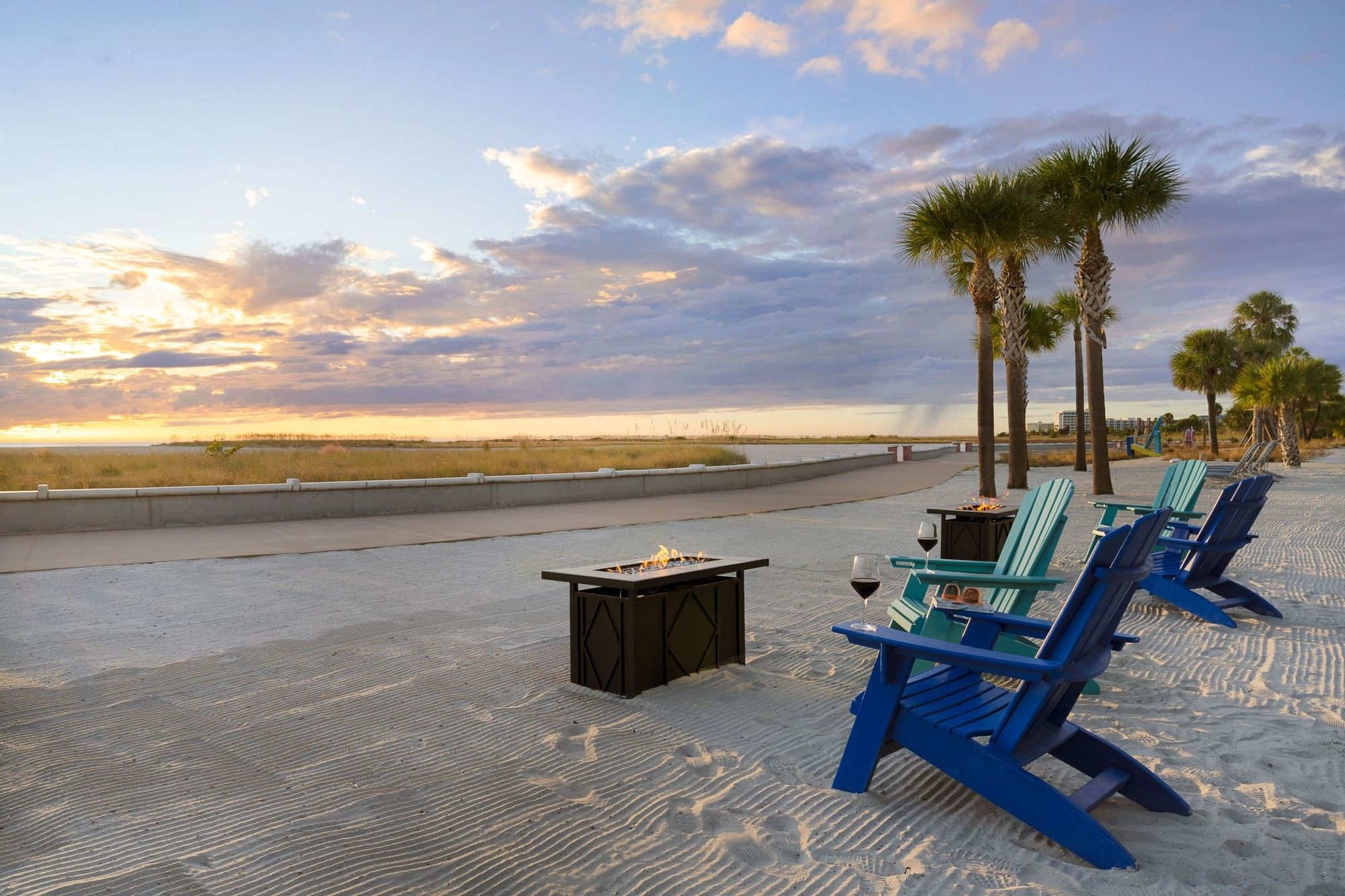 A beach scene with blue chairs, a fire pit, and palm trees at sunset.