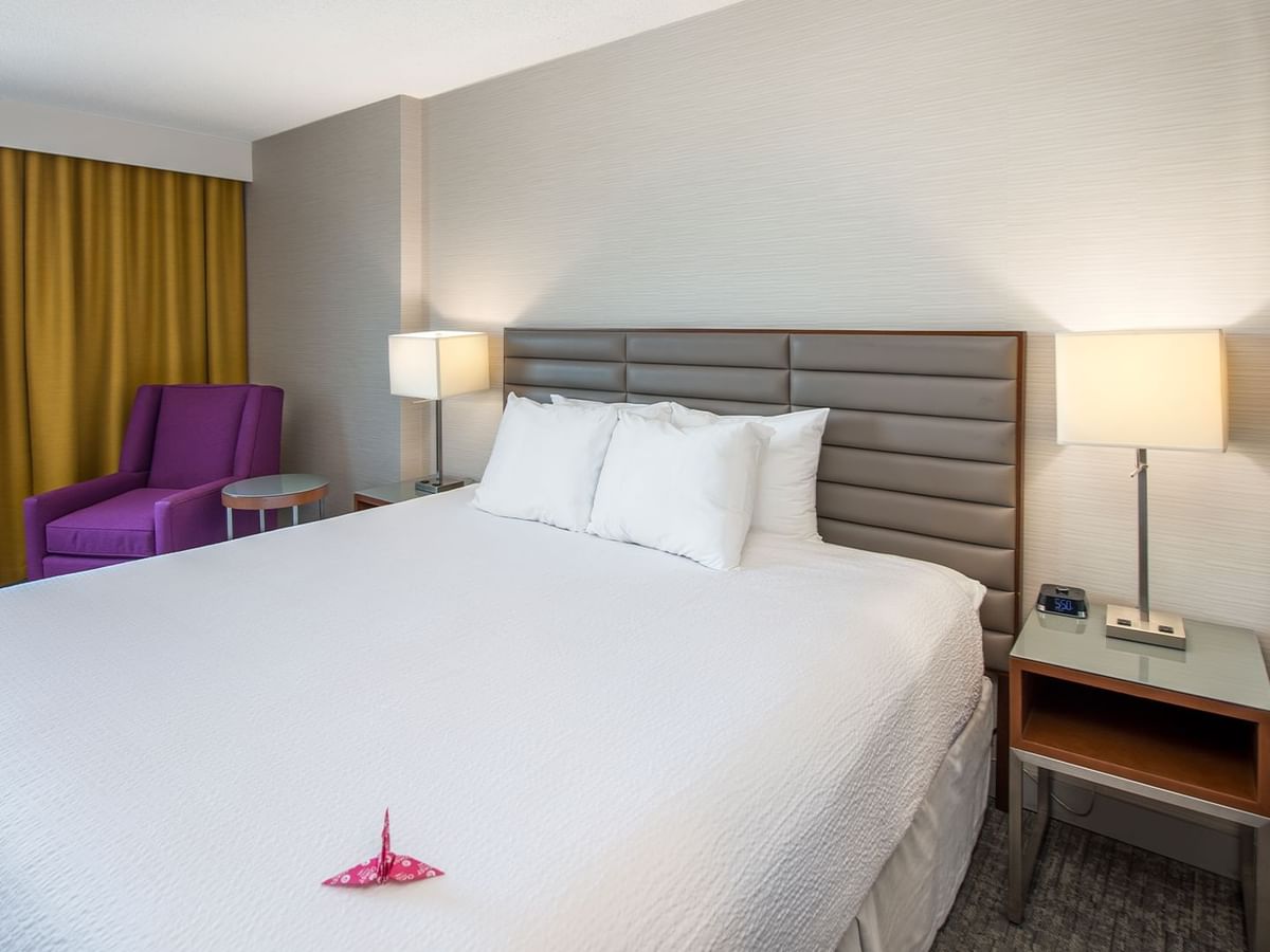 A hotel room with a white bed, two nightstands with lamps, a purple chair, and a yellow curtain.