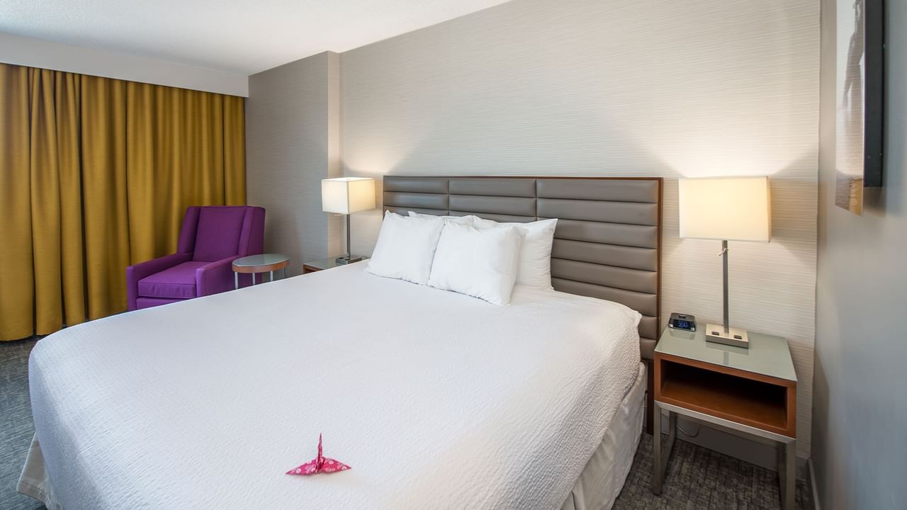A hotel room with a white bed, two nightstands with lamps, a purple chair, and a yellow curtain.