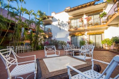 Outdoor seating area with white chairs and tables in front of a building with balconies and palm trees.