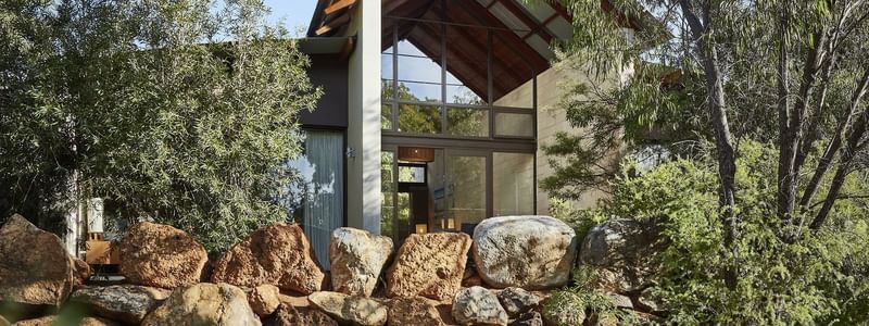 Modern glass-fronted accommodation nestled among native trees and natural stone landscaping under a clear blue sky.