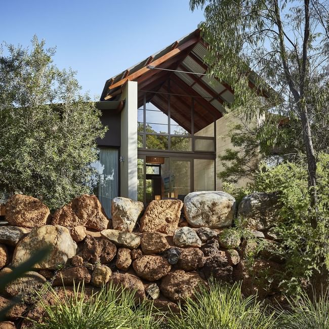 Modern glass-fronted accommodation nestled among native trees and natural stone landscaping under a clear blue sky.
