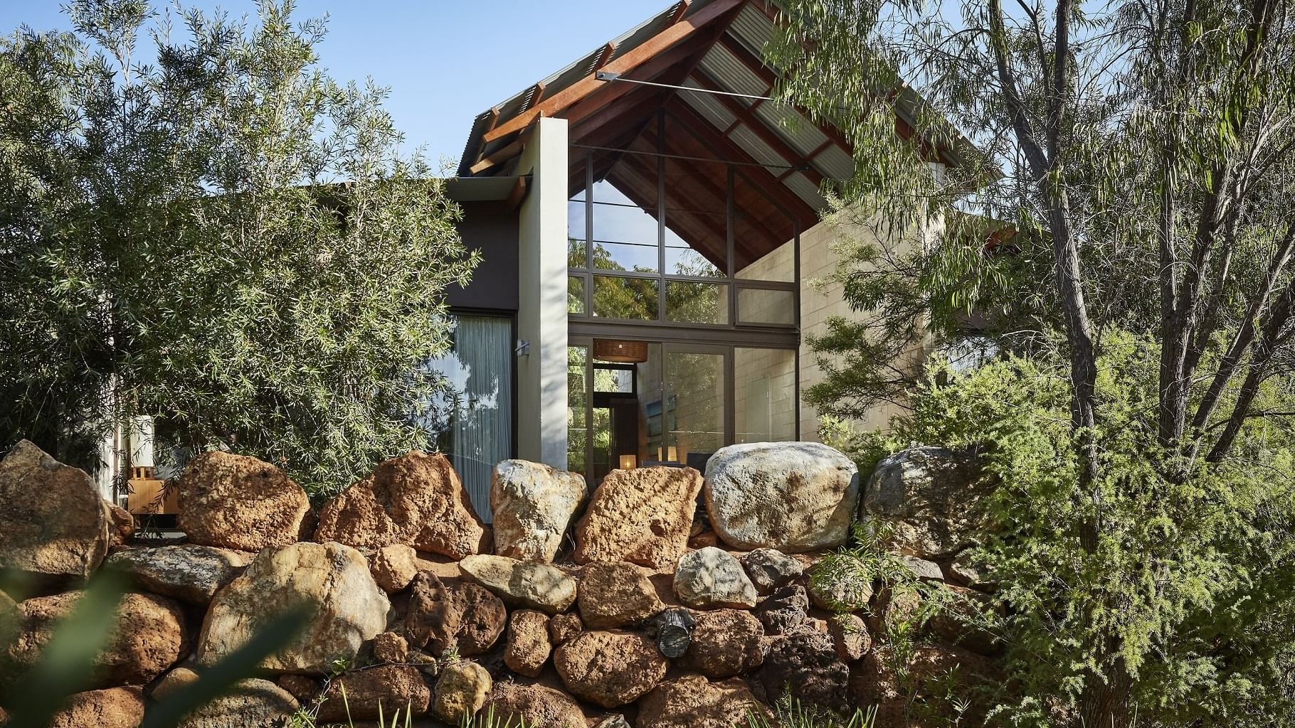 Modern glass-fronted accommodation nestled among native trees and natural stone landscaping under a clear blue sky.