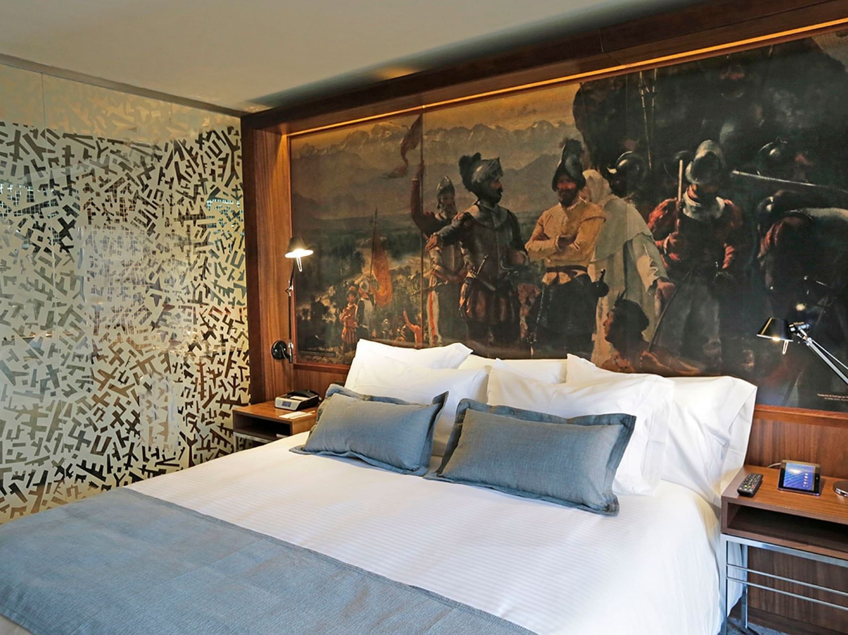 Large bed of the Standard Double room at the Cumbres Lastarria Hotel with blue pillows and blankets, and a large painting behind them.