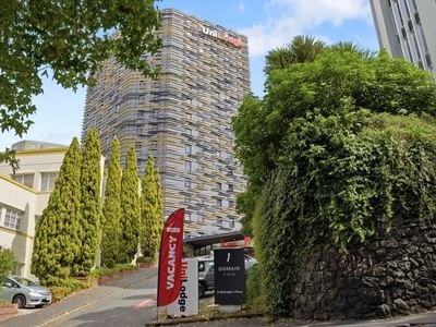 Tall UniLodge Auckland City building with trees and cars on the street in front.