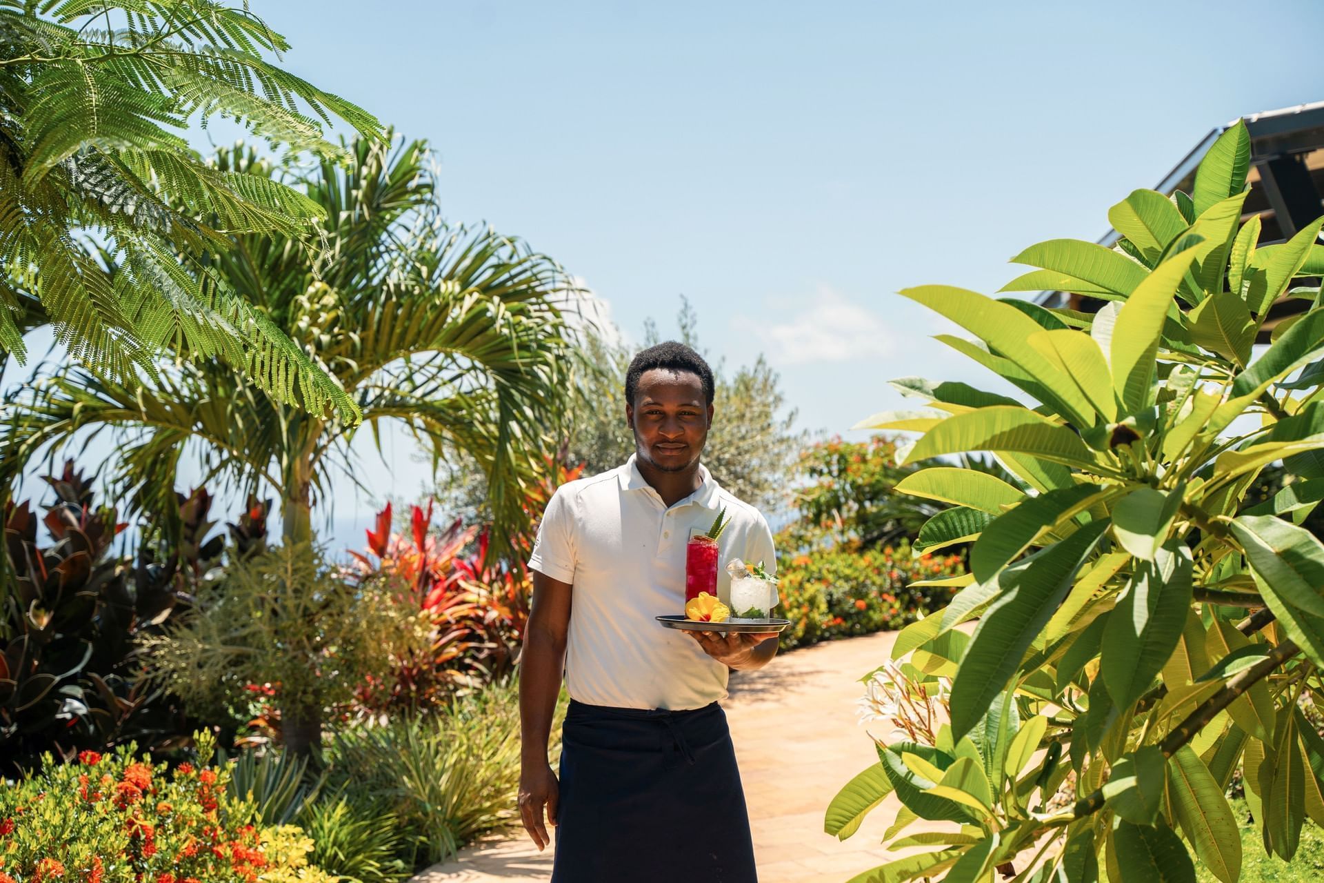 Ober met een dienblad met kleurrijke drankjes in de weelderige tuin van Breeze Restaurant bij Golden Rock Resort