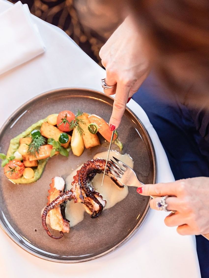Close-up of a guest slicing octopus, with roasted potatoes and tomatoes on a dark plate in Téséo at Warwick Geneva
