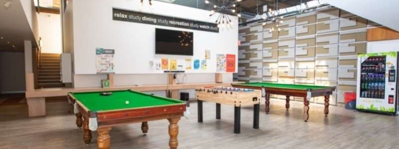 Two pool tables and one foosball table in a room with a wooden floor and a white wall.