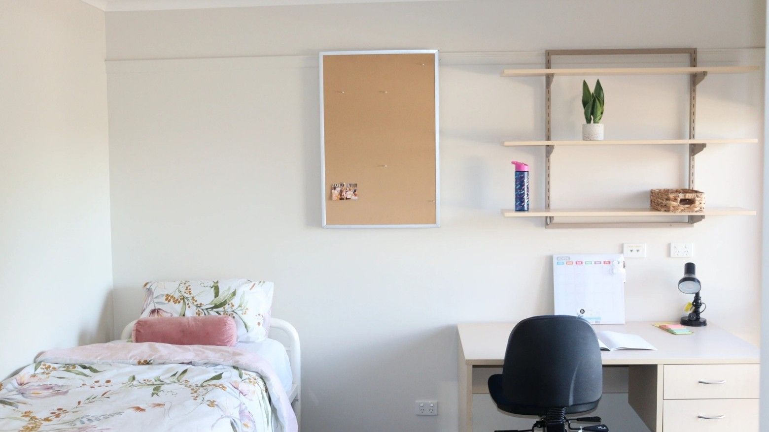 A room with a bed, desk, chair, bulletin board, shelves with items, and a calendar.