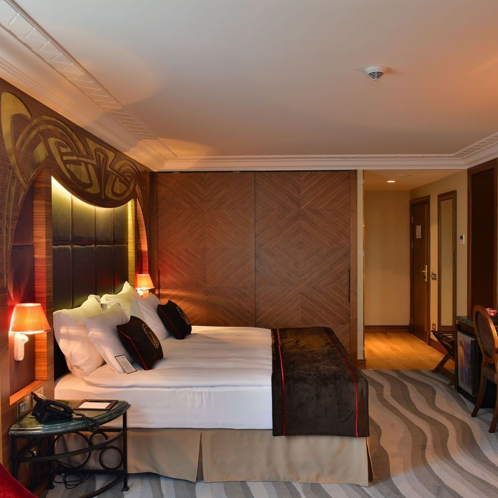 Work desk by a large bed with brown throws under a warm glowing light in Junior Suite at Warwick Ankara