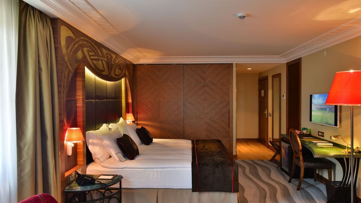 Work desk by a large bed with brown throws under a warm glowing light in Junior Suite at Warwick Ankara