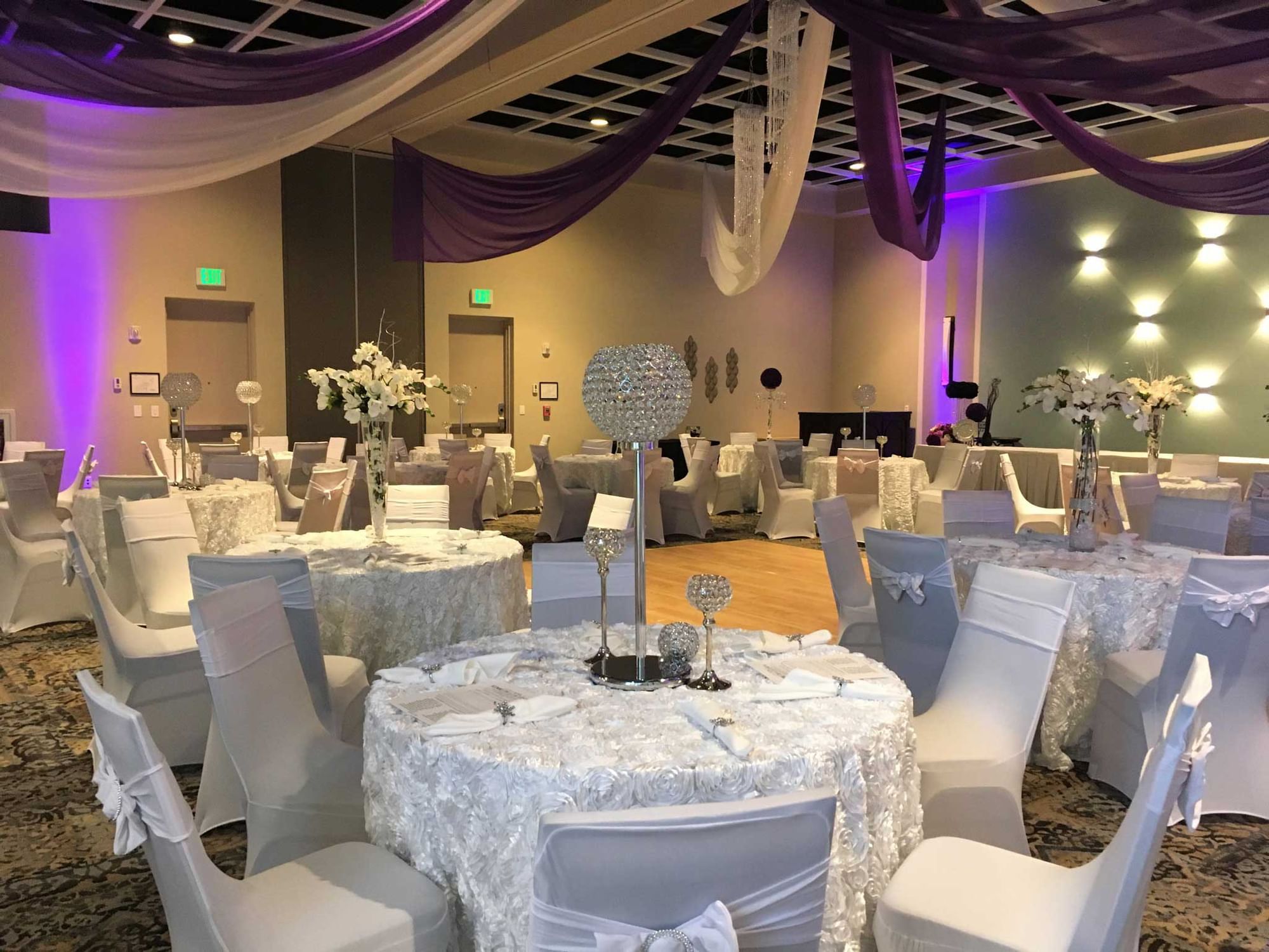 A large banquet hall with round tables set for a formal event, featuring white chairs, purple drapes, and floral arrangements.