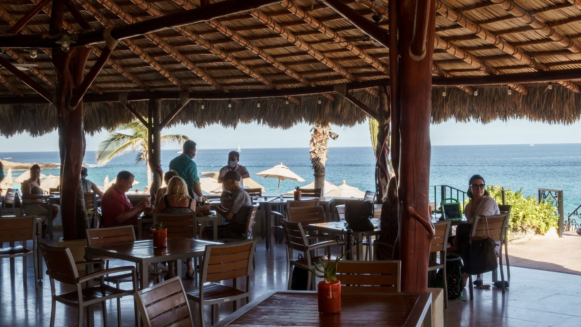 Diners at tables under a thatched roof with a beautiful ocean view at Sirenas Restaurant in Hacienda Del Mar Los Cabos.