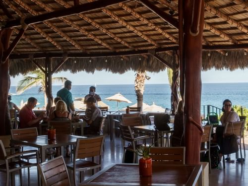 Diners at tables under a thatched roof with a beautiful ocean view at Sirenas Restaurant in Hacienda Del Mar Los Cabos