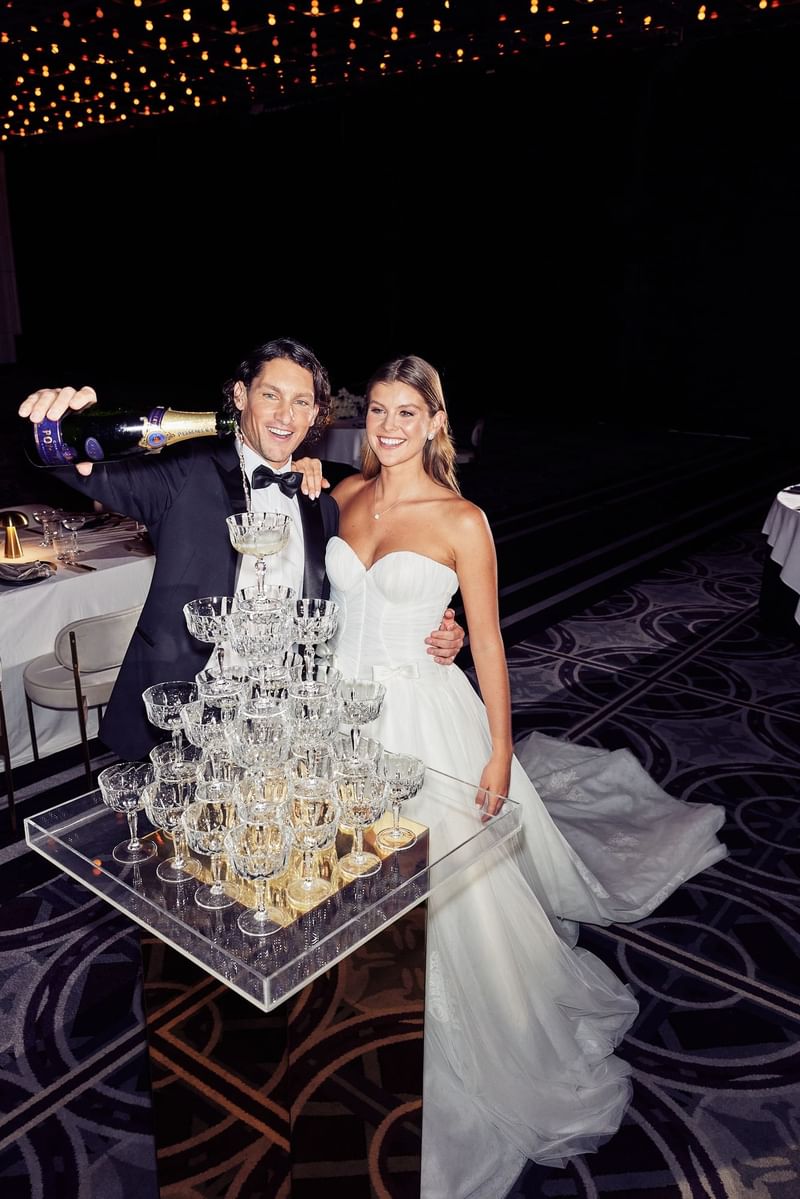 A groom pours champagne into a pyramid of glasses as his bride stands by his side.