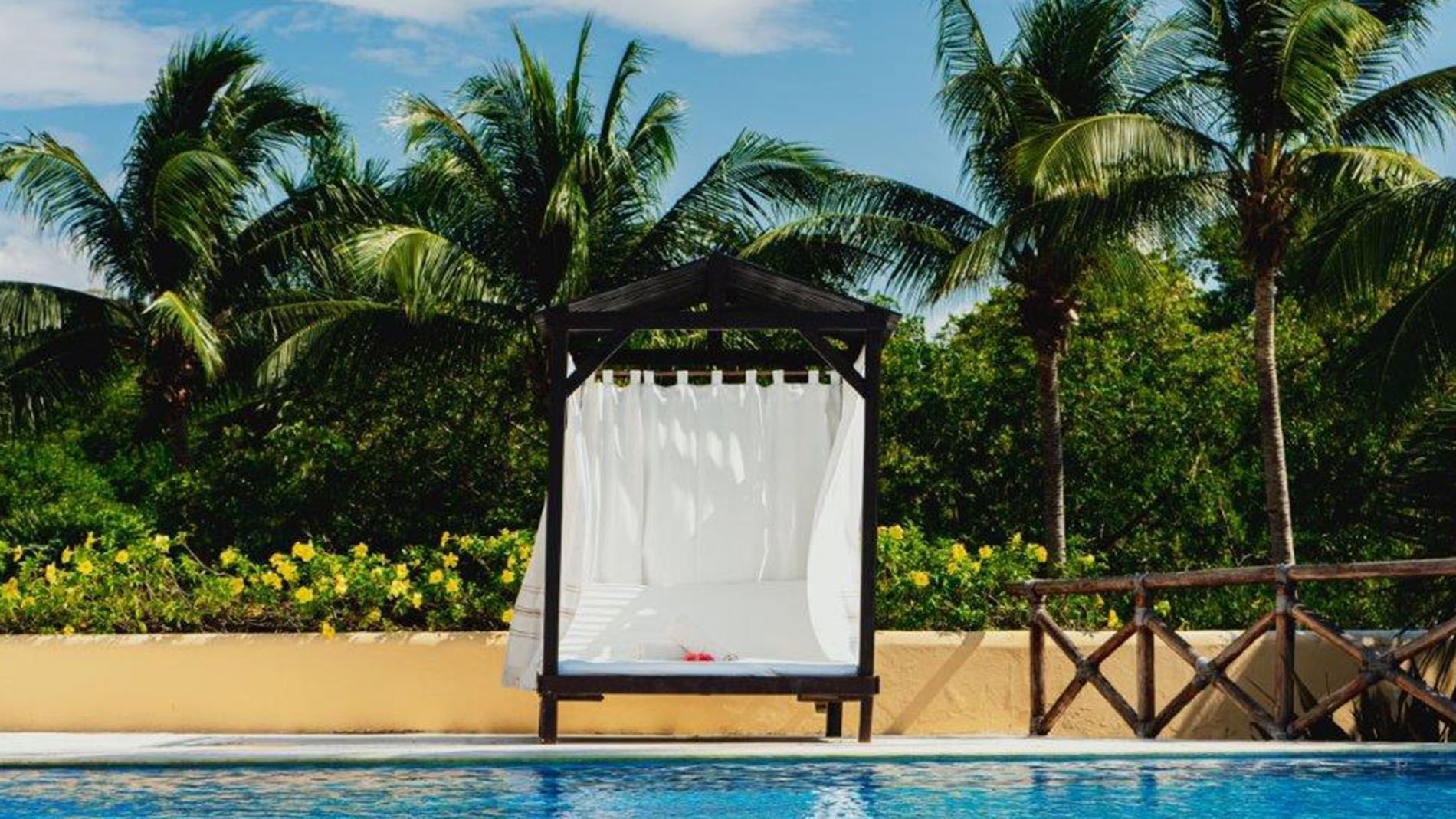 Poolside cabana with white curtains by a pool under palm trees surrounding a tropical garden at Quinta Real Huatulco