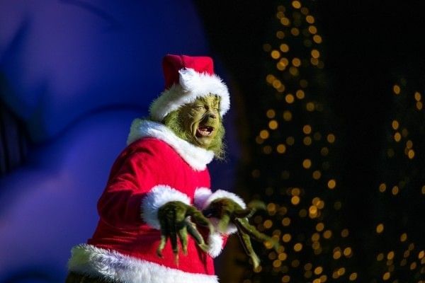 The Grinch in a Santa suit standing onstage in front of a blurred Christmas tree during the holidays at Universal Orlando.