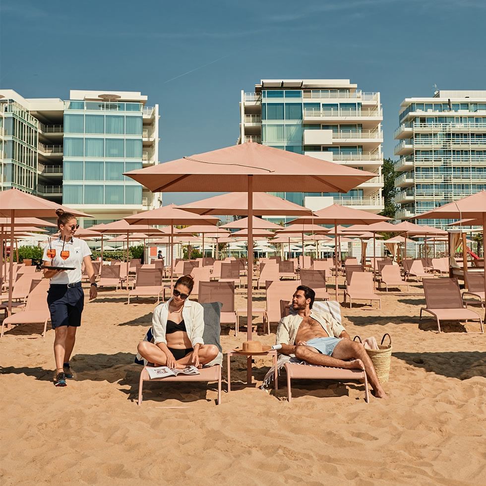 People relaxing on beach lounge chairs under umbrellas with buildings in the background at Falkensteiner Hotel & Spa Jesolo