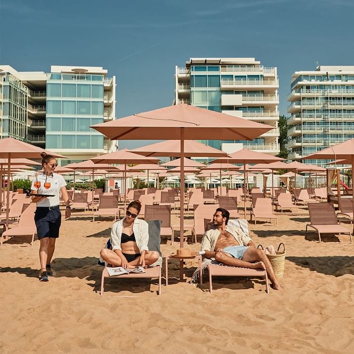 Gäste am Strand mit Liegen und Sonnenschirmen vor Gebäuden an einem sonnigen Tag.
