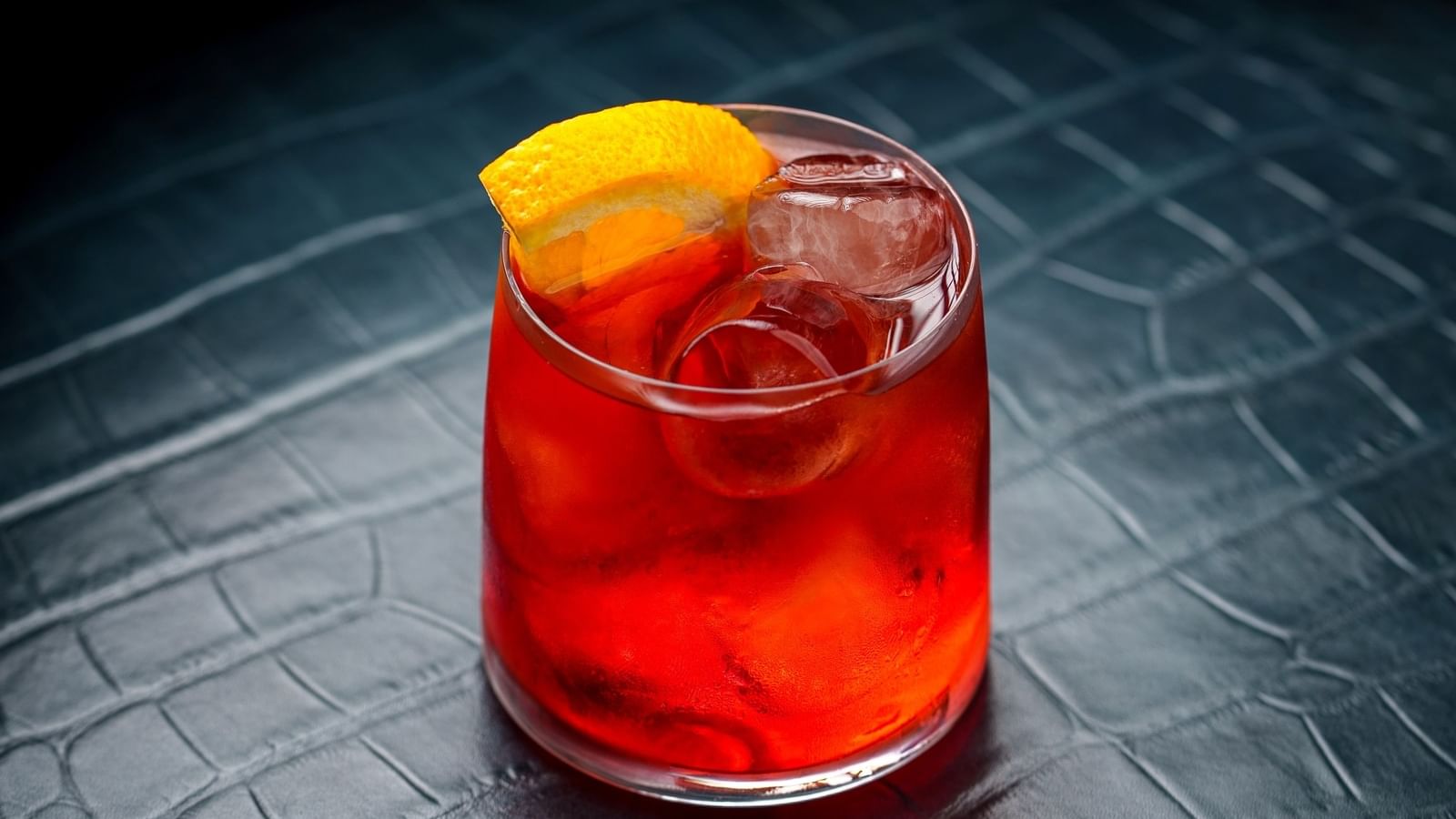A red cocktail with ice and a lemon slice in a clear glass on a black leather surface at Warwick Hotels and Resorts