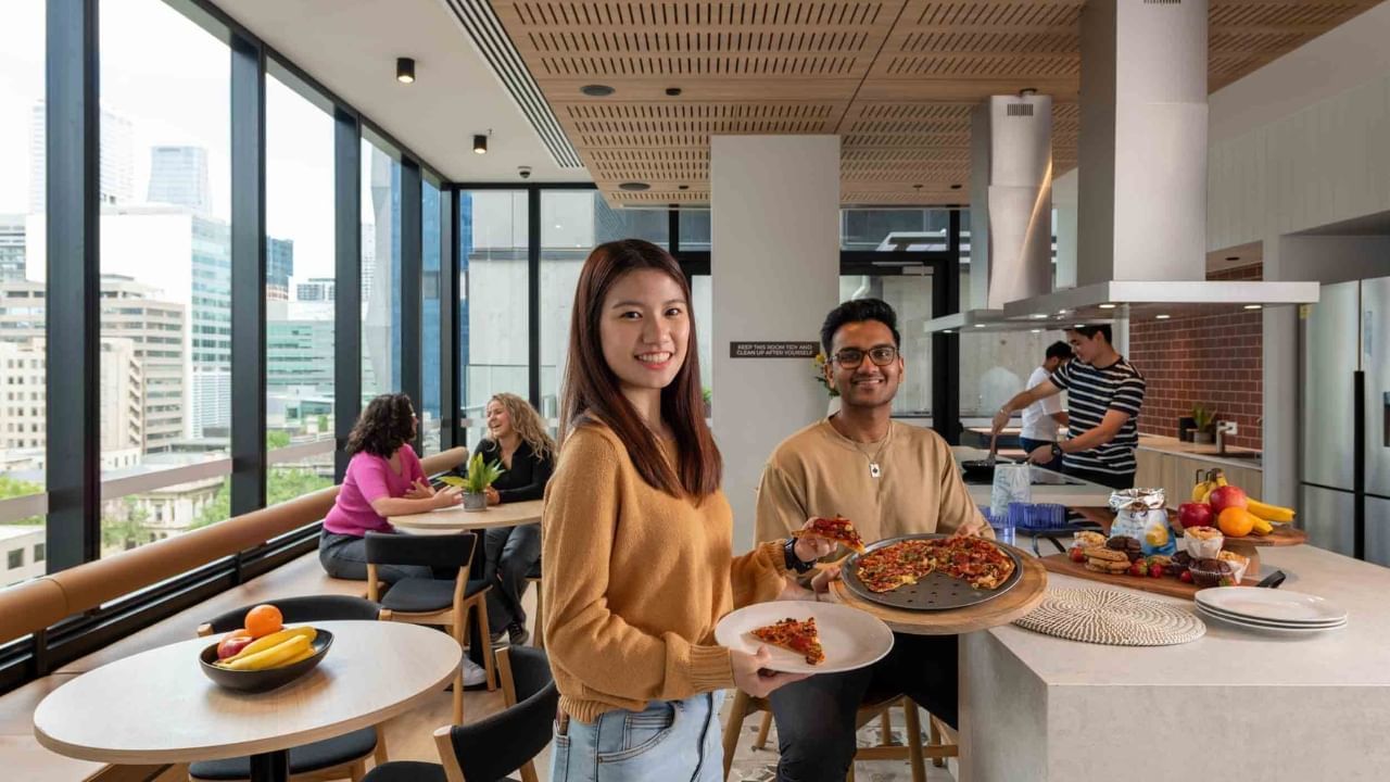 Residents holding pizza plates smile at the camera in a communal kitchen.