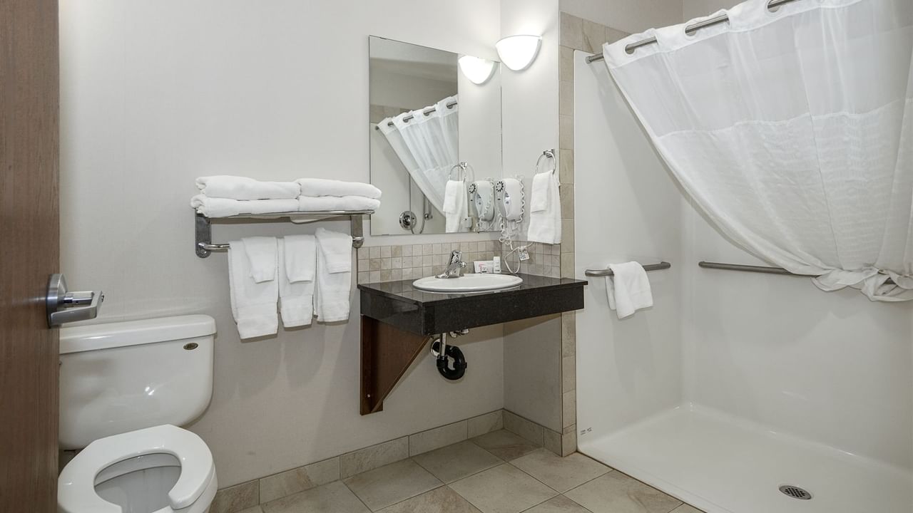 Accessible hotel washroom with roll-in shower, toilet, sink, towels and soap