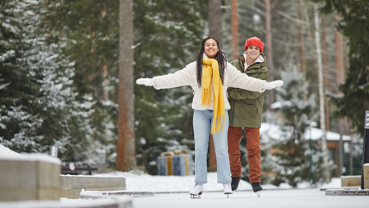 Two smiling ice skaters with arms outstretched glide together surrounded by snowy pines.