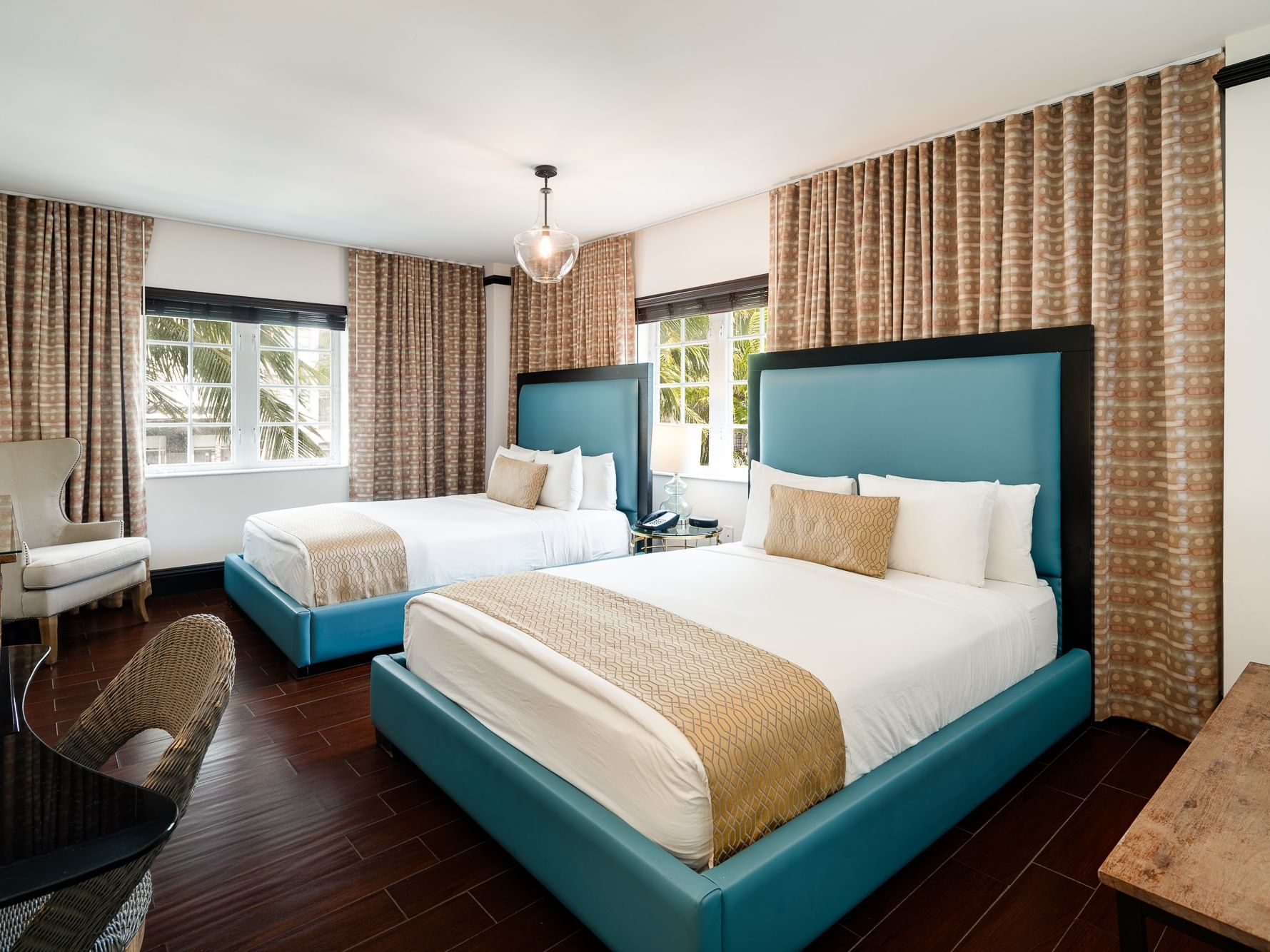 Two queen beds with blue headboards and white bedding, wooden flooring, and two chairs near windows with curtains.